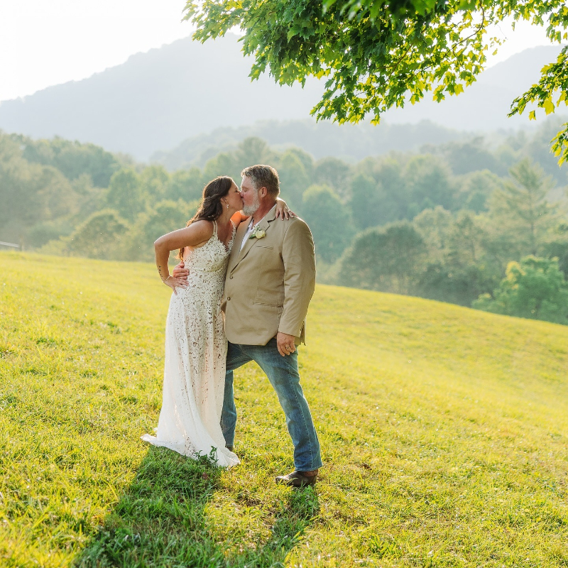 Couple shares a kiss in a sunny mountain meadow, surrounded by trees and rolling hills.