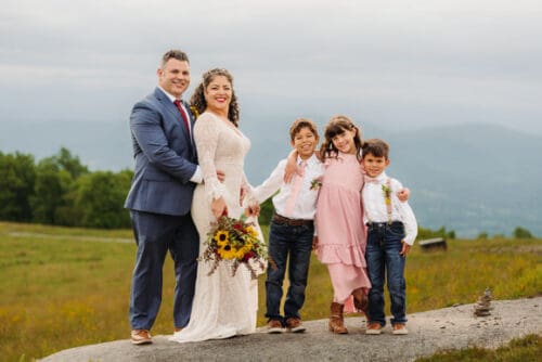 Smiling couple with three children pose on a mountaintop, blending family joy with scenic beauty.