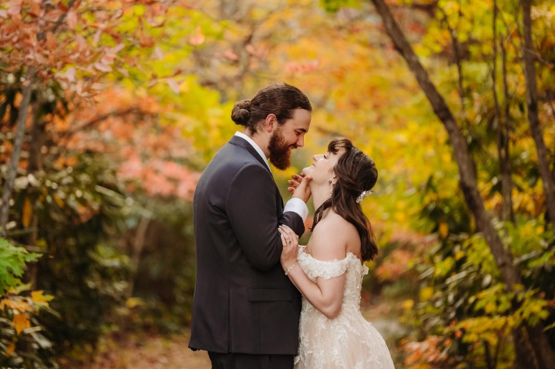 Bride and groom share a tender moment surrounded by vibrant fall leaves in a forest path.
