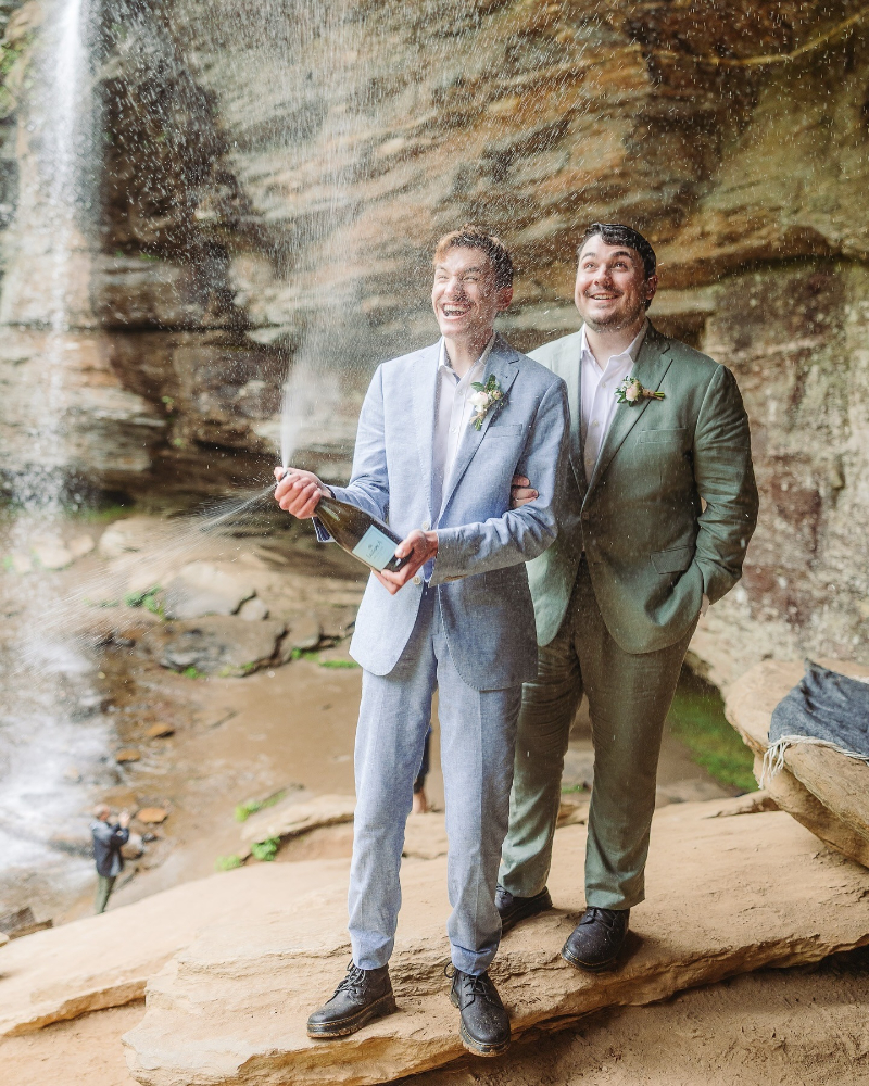Two grooms celebrate with champagne during joyful elopement planning at a waterfall in Asheville.