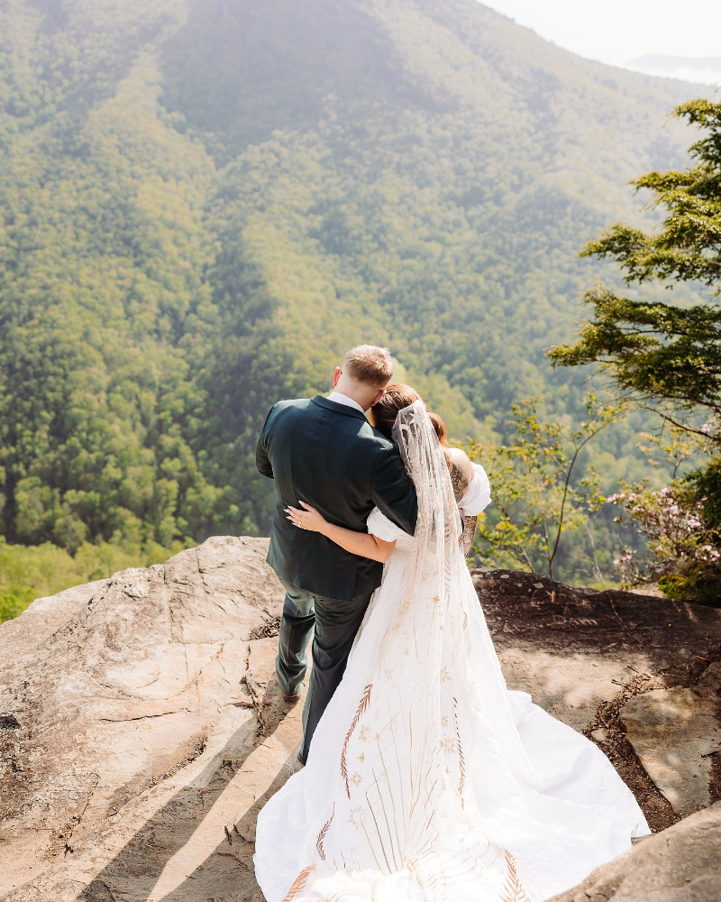 Couple embraces on mountain cliff during elopement planning, overlooking lush Blue Ridge forest views.
