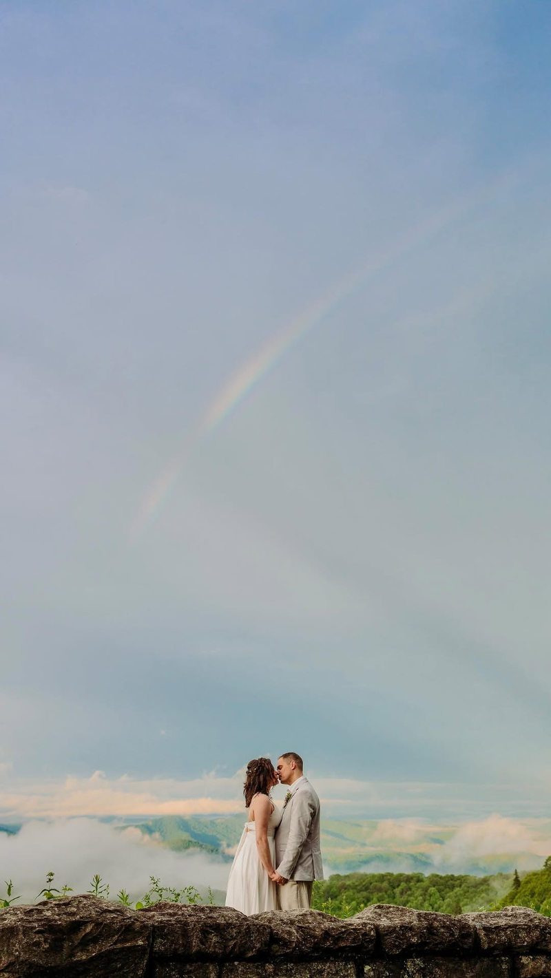 Couple kisses during elopement planning in the Blue Ridge Mountains beneath a soft rainbow sky.