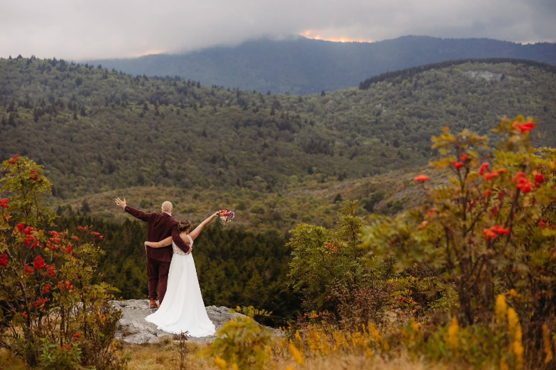 Couple stands on a mountain ledge with arms raised, overlooking rolling hills at sunset.