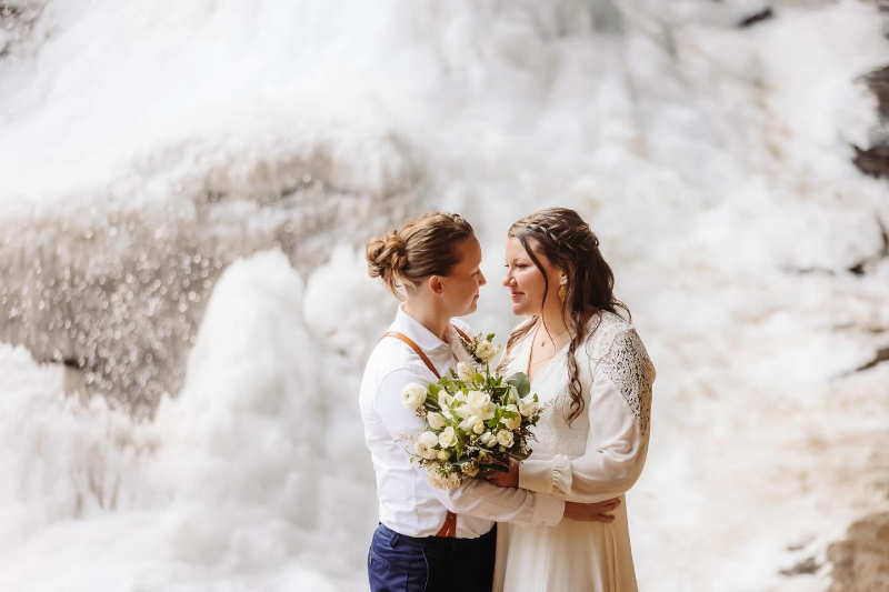 Couple stands close with a white bouquet in front of a frozen waterfall during their winter elopement.