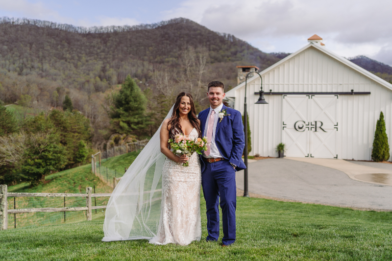 Bride and groom smiling on green lawn with white barn and mountain backdrop.
