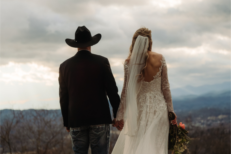 Bride and groom holding hands, overlooking layered mountains at sunset.