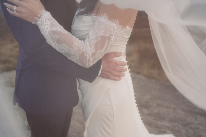 Close-up of couple embracing during intimate elopement ceremony, bride in lace sleeve, groom holding her waist outdoors.