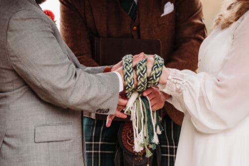 Couple performing handfasting ritual during outdoor elopement ceremony, colorful cords tied around their hands.