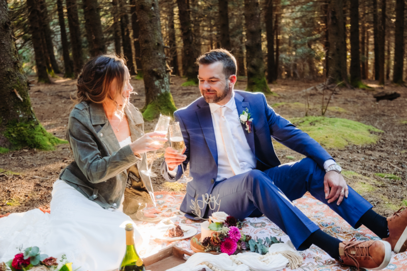 Couple enjoying a champagne picnic together on a blanket in a grassy field with flowers and glasses.