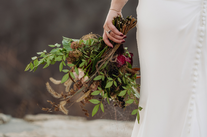 Bride holding a natural bouquet with greenery and dried florals beside a simple wedding dress.