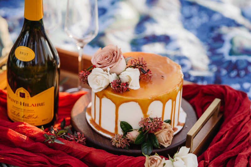 Small wedding cake with caramel drizzle and flowers beside a bottle of champagne.