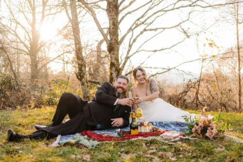 Couple relaxing on a picnic blanket with champagne and cake during a sunset celebration outdoors.