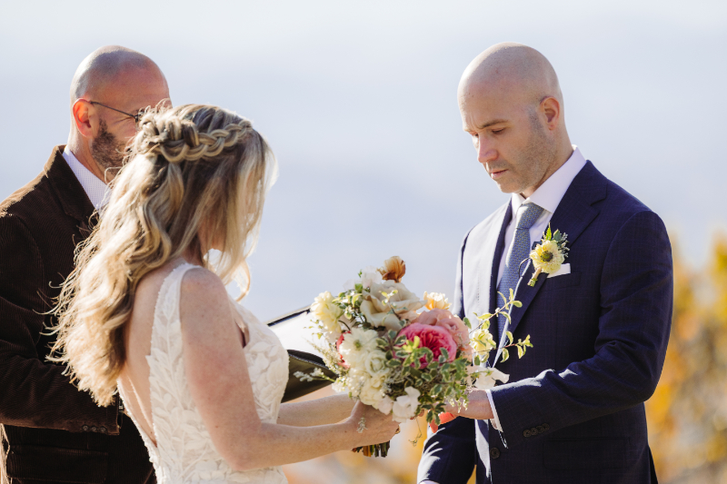 Bride and groom exchanging vows, holding hands with bouquet in a soft mountain setting