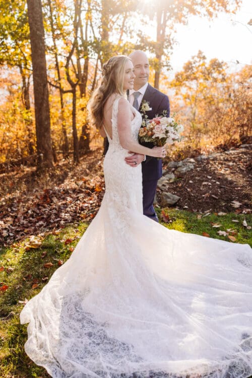 Bride standing in a forest with golden fall leaves, holding bouquet and wearing a flowing dress