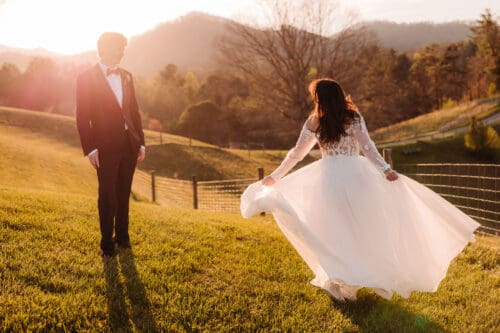 Bride twirling in a flowing lace wedding dress at sunset while groom watches in a golden mountain field in the Blue Ridge Mountains
