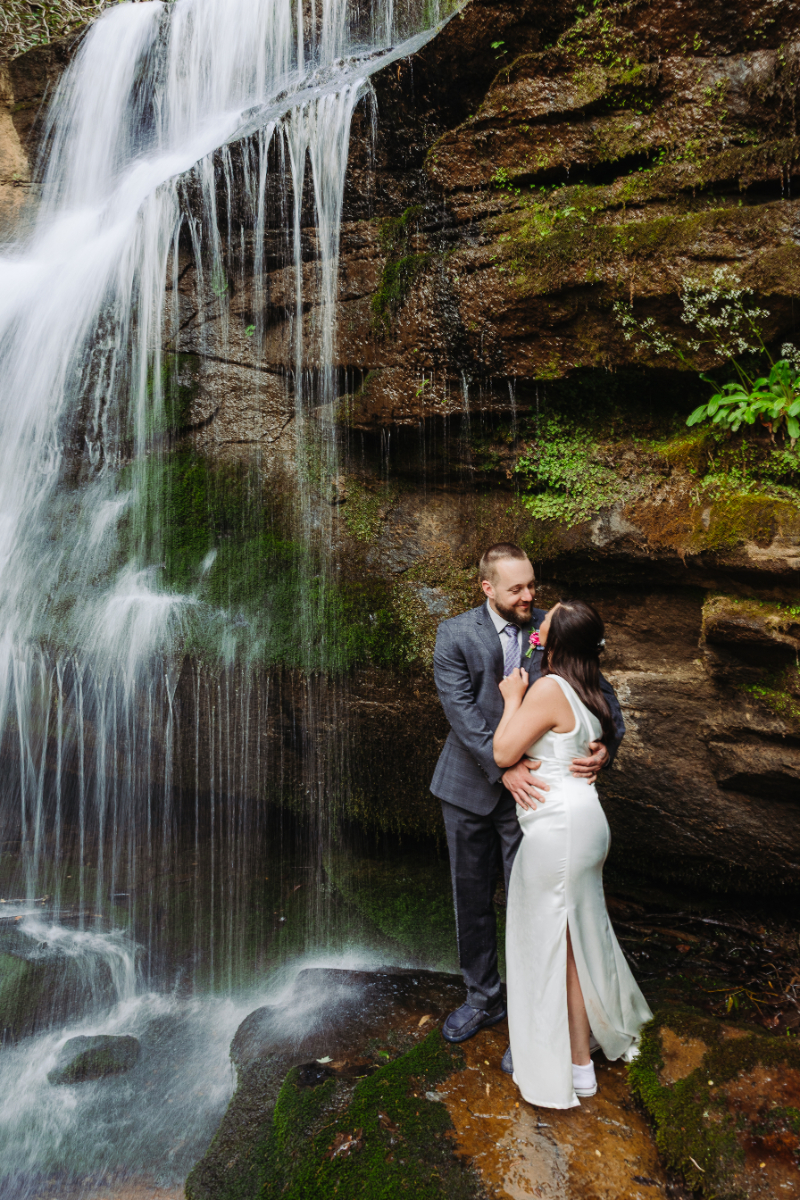 Couple embracing beside a cascading waterfall, surrounded by mossy rocks and soft flowing water