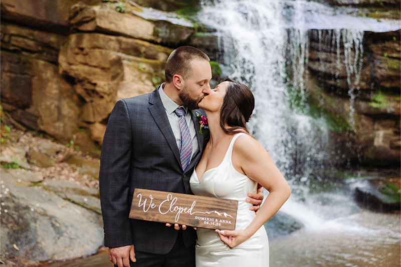 Couple kissing by a waterfall, holding a just married sign with flowing water behind them