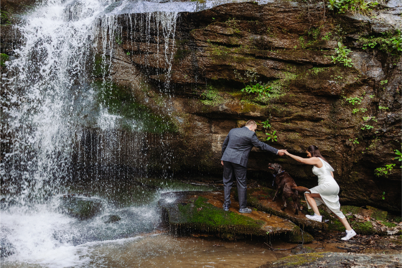 Couple helping each other across wet rocks beside a flowing waterfall in a forest setting