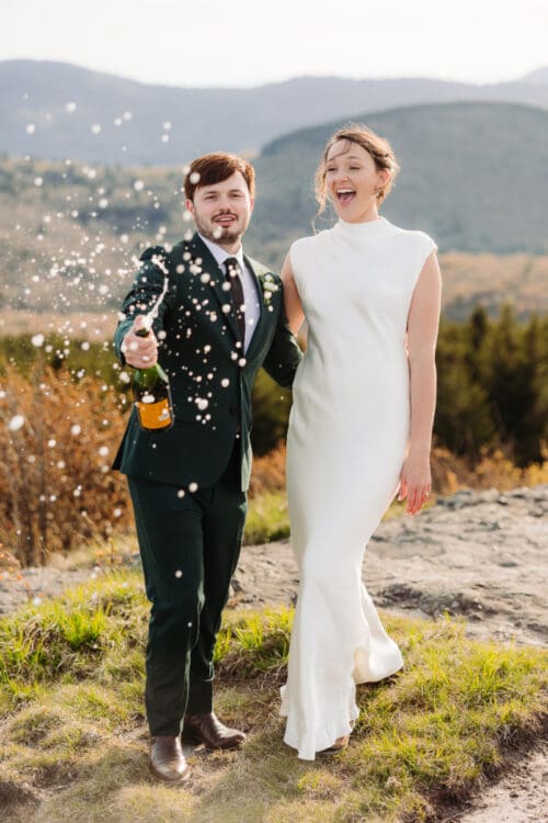 Newlyweds celebrating outdoors while spraying champagne with mountain views in the background.