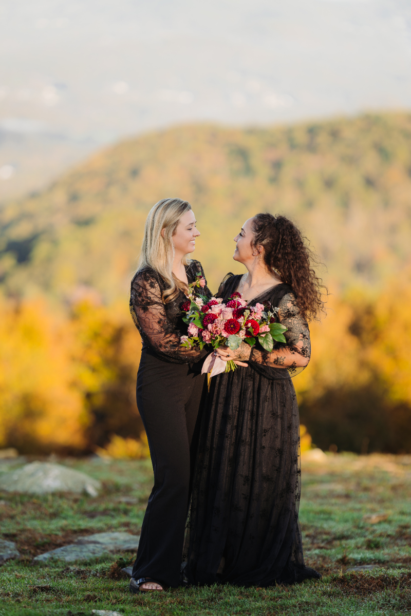 Couple holding bouquets and smiling at each other with mountain views and autumn foliage.