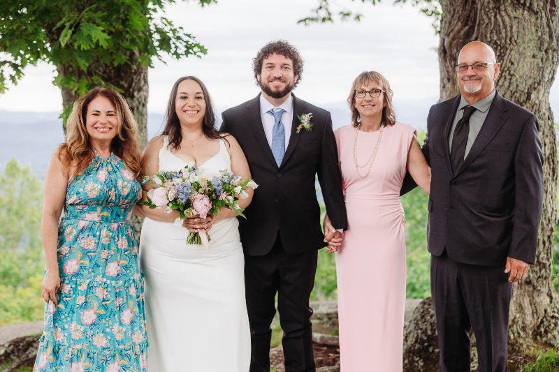 Bride and groom pose with family members after their mountain wedding ceremony beneath large trees.