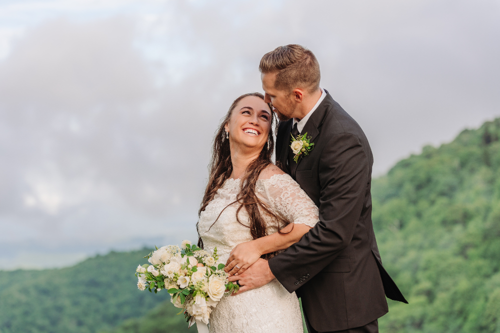 Couple embracing with a bridal bouquet against a peaceful Blue Ridge Mountain backdrop after their ceremony.