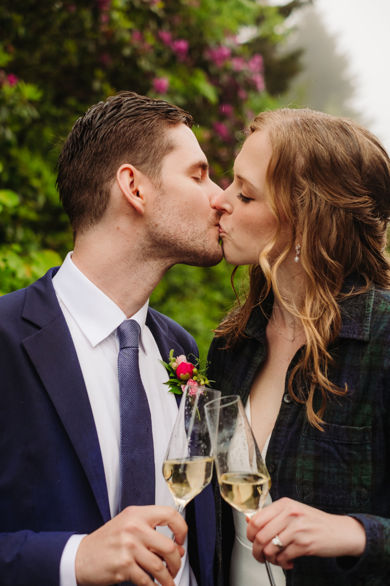 Newlyweds sharing a kiss while holding champagne glasses after their forest elopement ceremony.