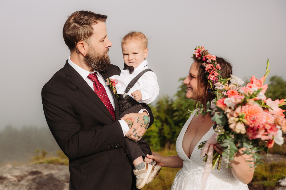 Bride and groom smile at their young child during a family focused mountain wedding.
