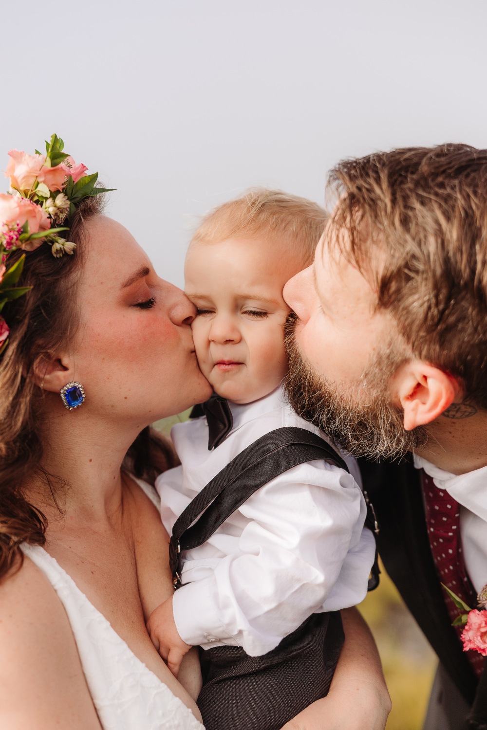 Bride and groom kiss their young child during a joyful mountain wedding celebration.