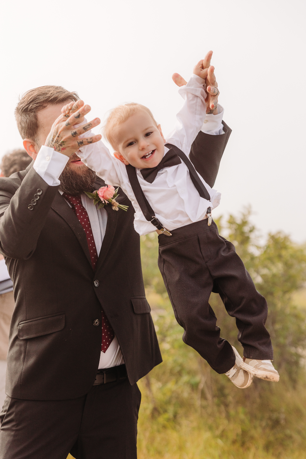 Groom lifts young child into the air during an outdoor mountain wedding.