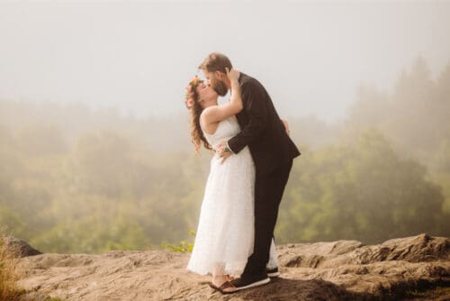 Bride and groom share a kiss on a scenic mountain overlook at sunset.