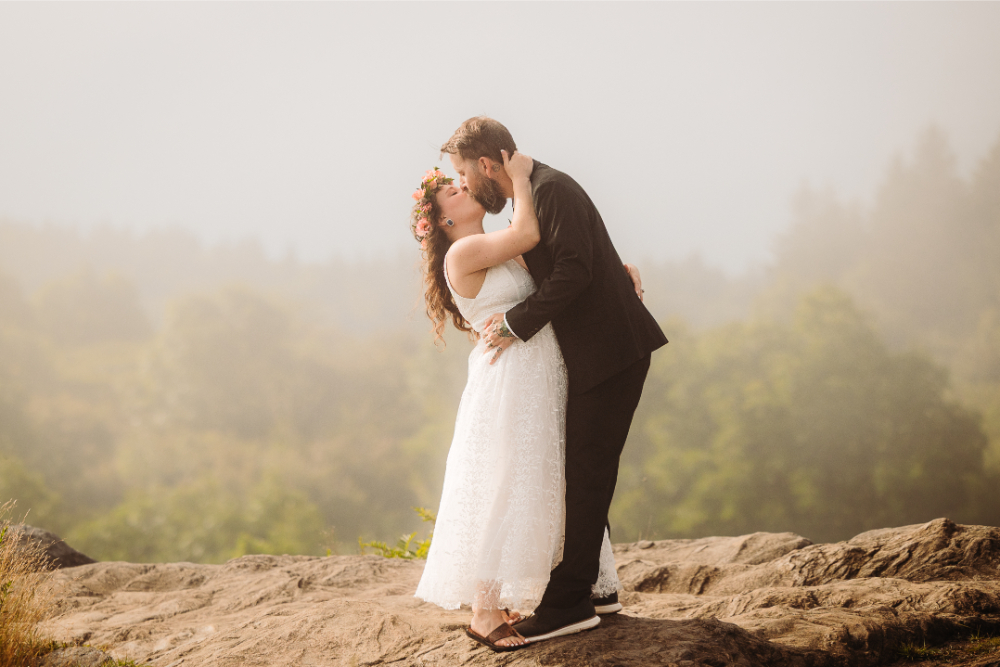 Bride and groom share a kiss on a scenic mountain overlook at sunset.