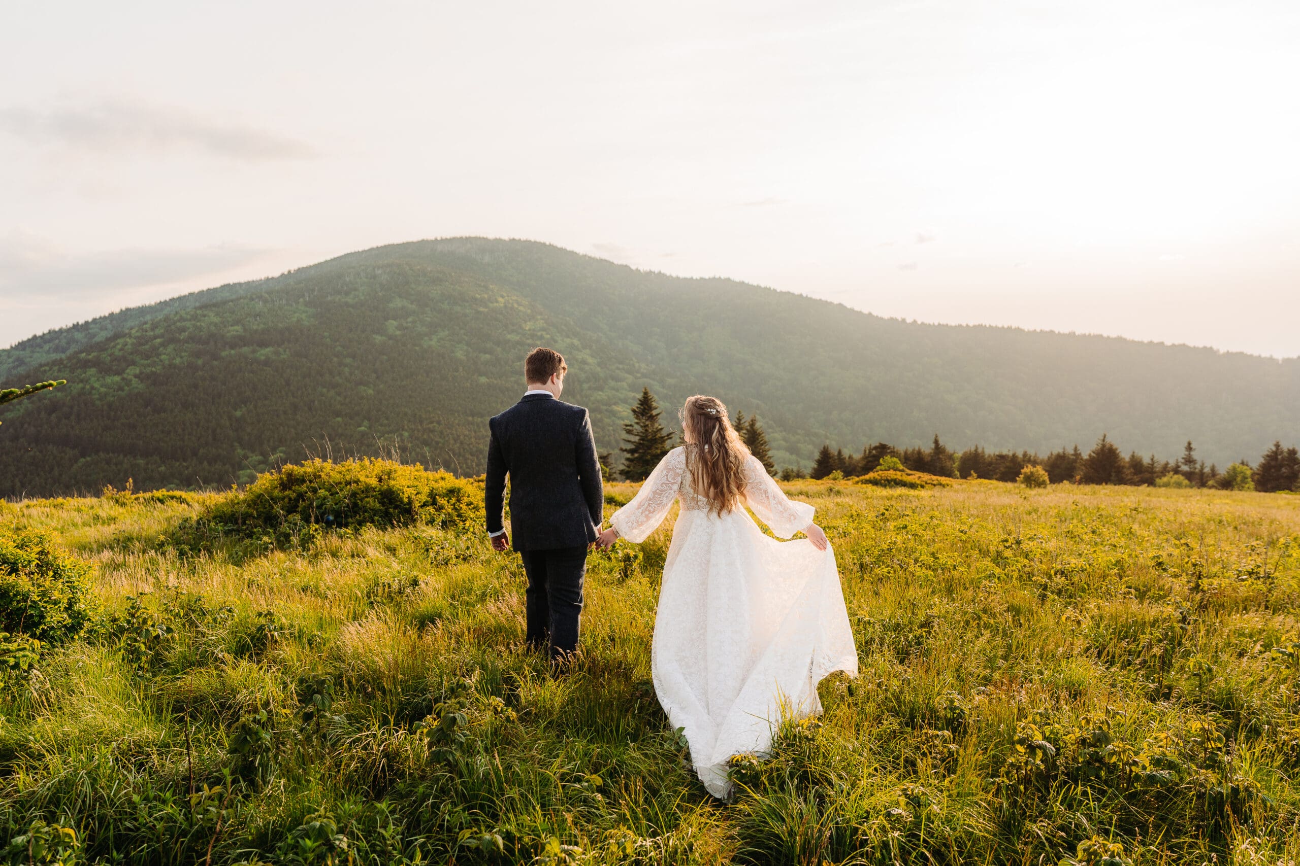 Newlyweds walking hand in hand through a grassy Blue Ridge Mountain meadow at sunset