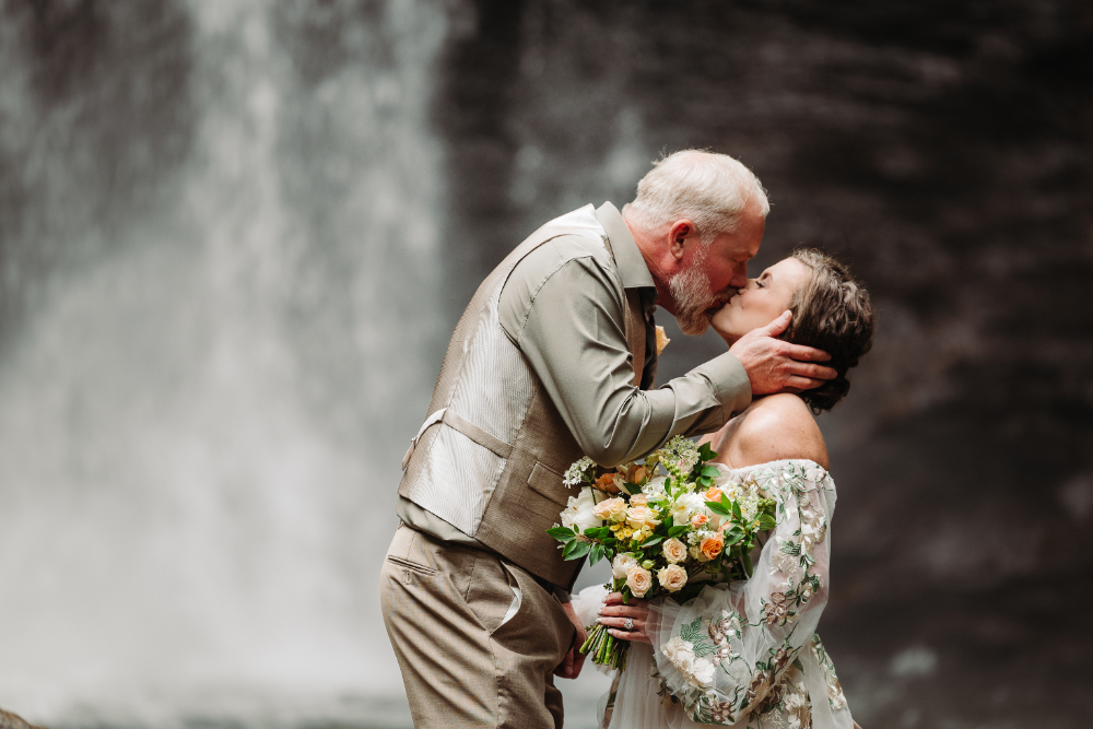 Newlywed couple sharing a romantic kiss in front of a beautiful waterfall during their outdoor elopement.