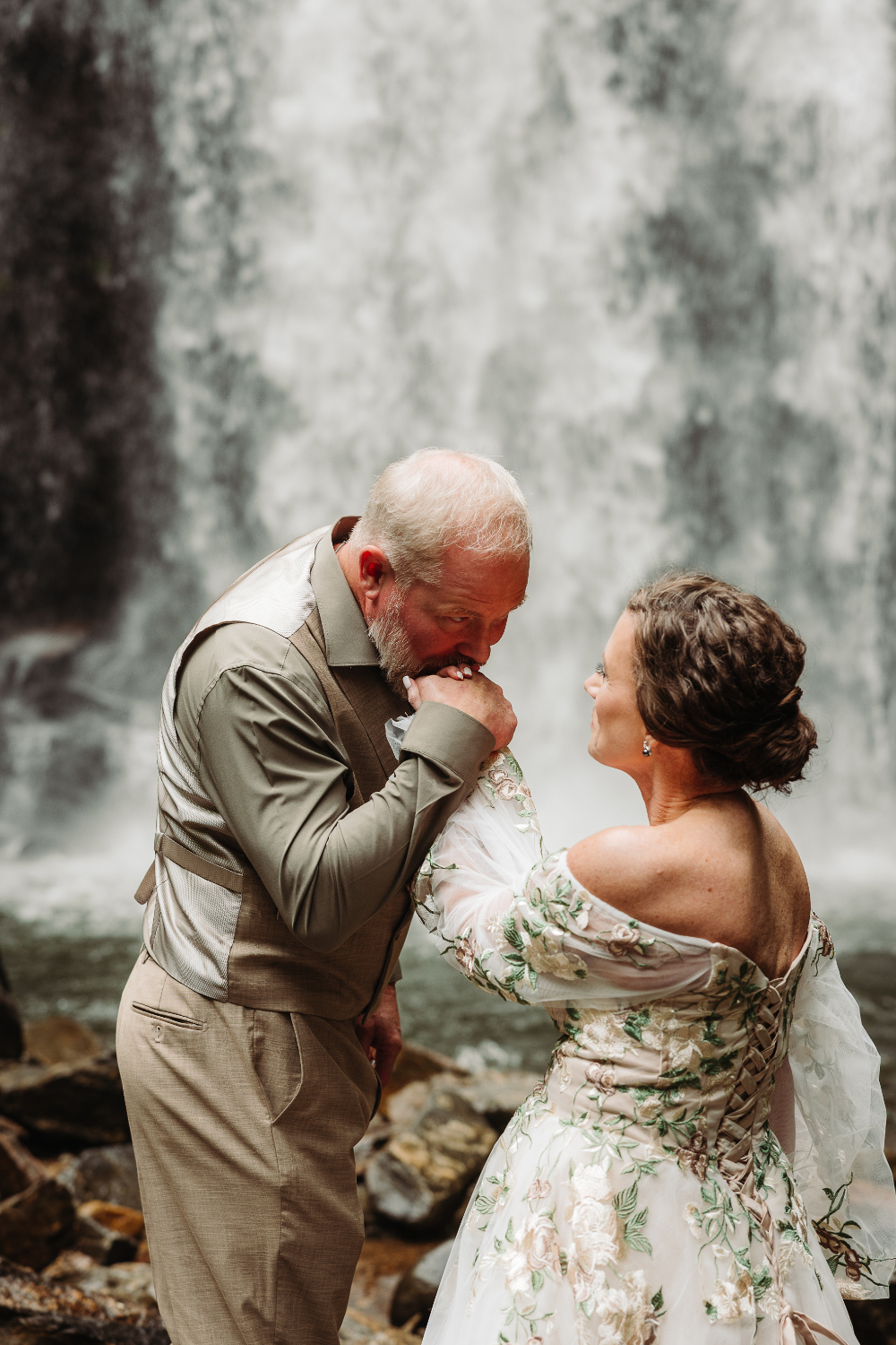 Groom kissing his bride’s hand beside a waterfall during their intimate wedding over 40.