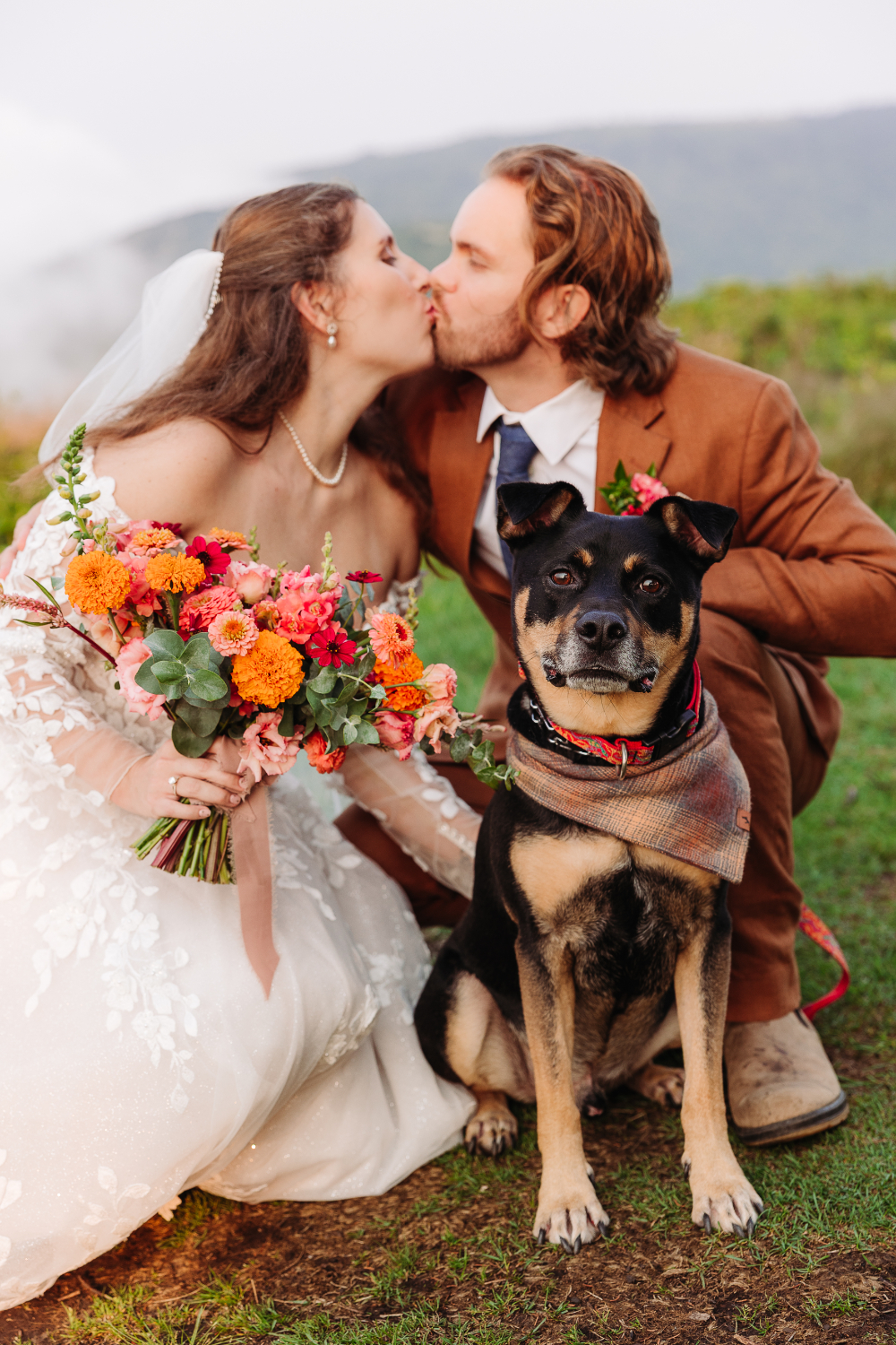 Couple kissing with their dog during an Asheville elopement, holding a colorful bridal bouquet.