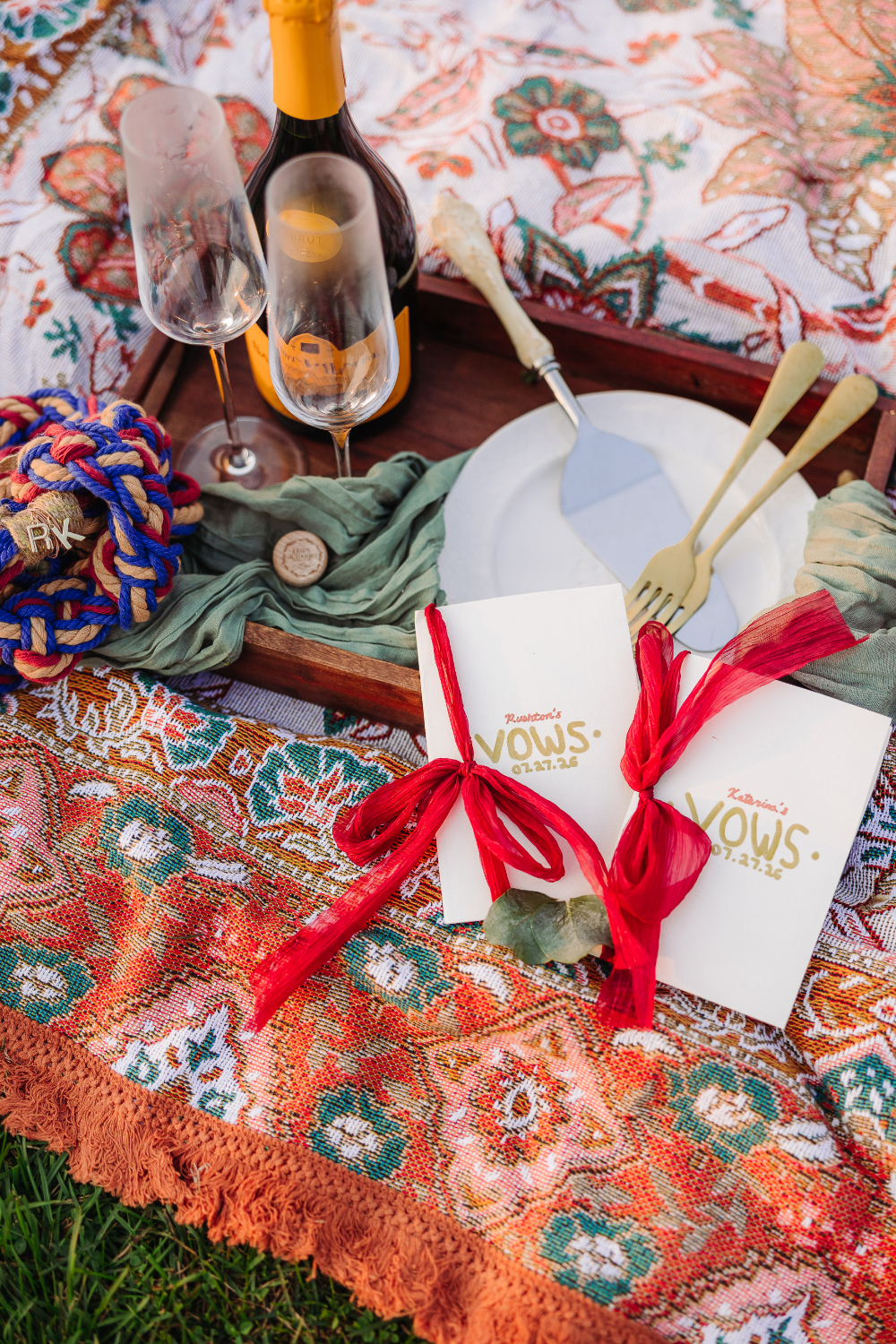 Personalized vow books, champagne glasses, and keepsakes arranged on a colorful picnic blanket.