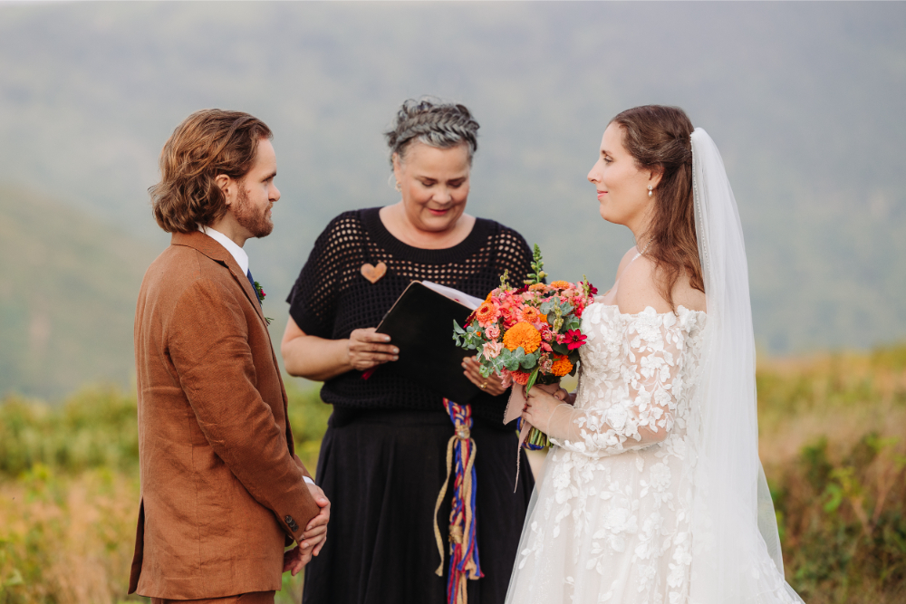Asheville wedding officiant smiles with a couple during their relaxed outdoor ceremony overlooking the Blue Ridge Mountains.