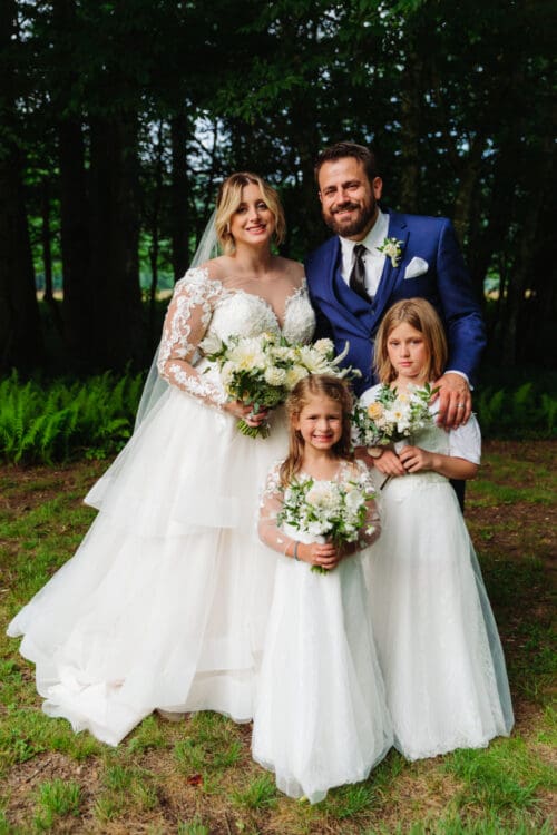 Newlywed couple posing with two young girls holding white flower bouquets in the forest.