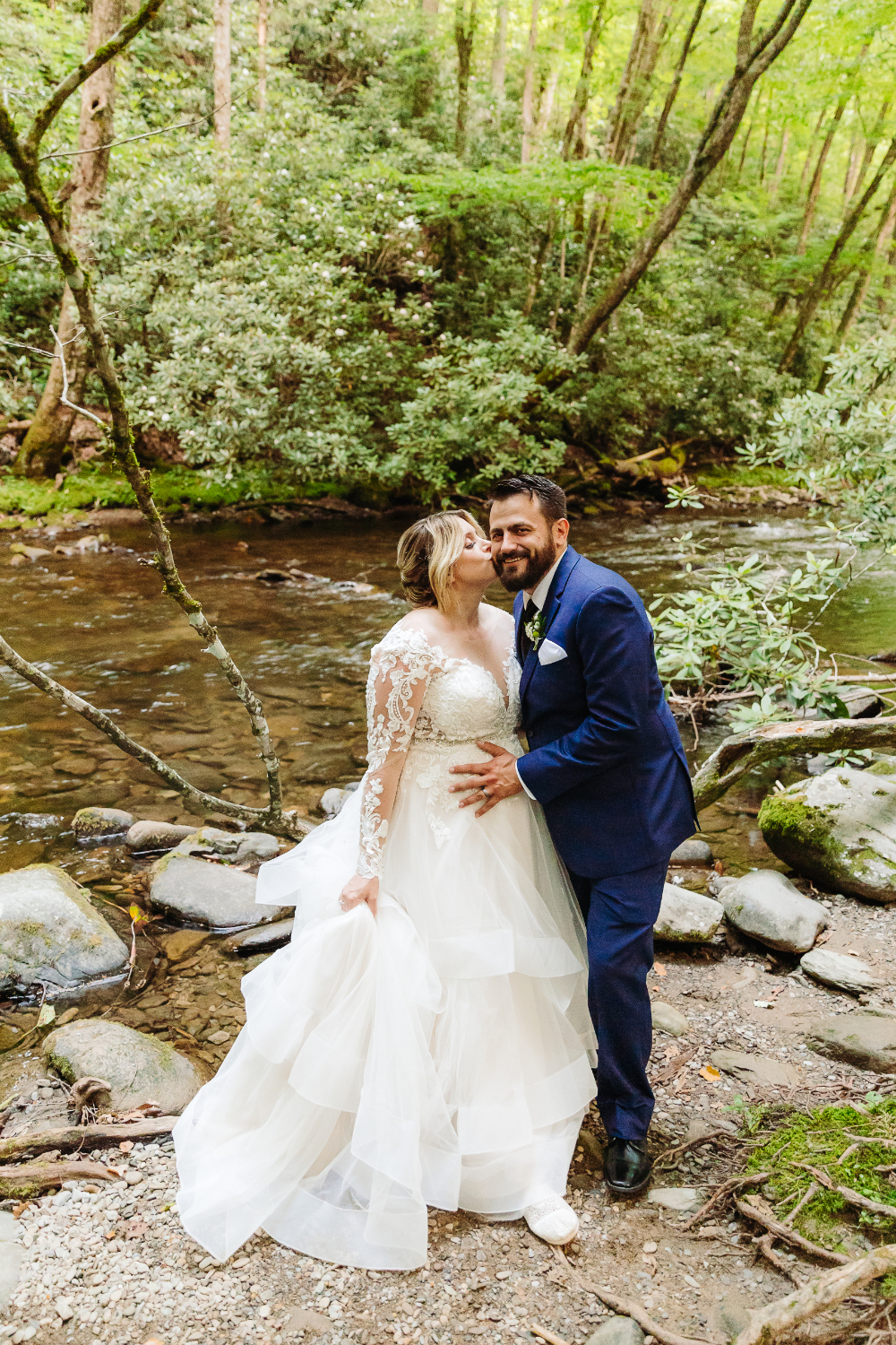 Bride and groom embracing beside a peaceful rocky stream surrounded by lush forest.