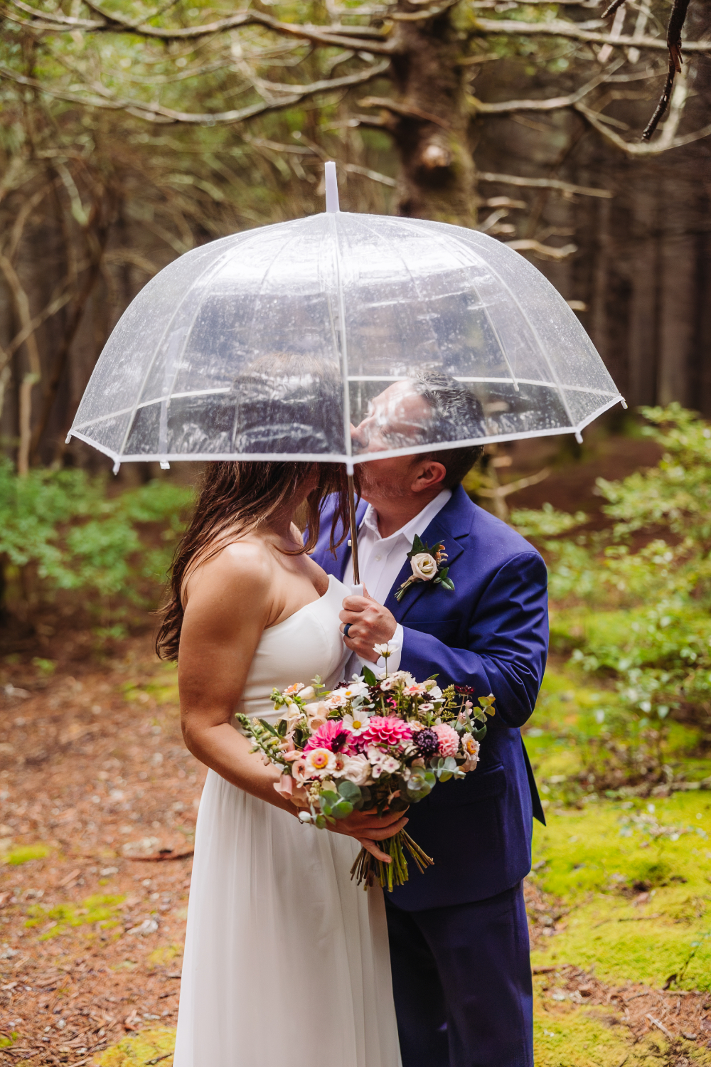 Bride and groom sharing a kiss beneath a clear umbrella in the rain, inspiring romantic intimate wedding ideas.